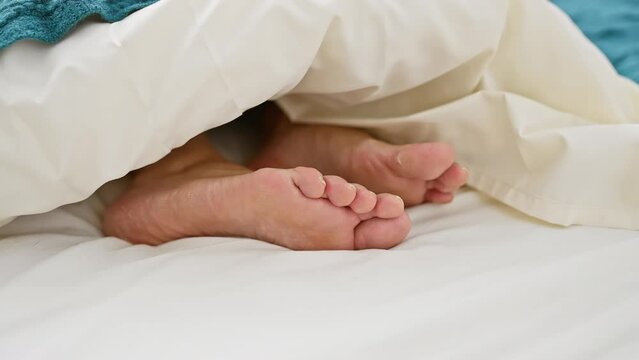Close-up image of adult bare feet under white bed sheets, suggesting relaxation or sleep in a bedroom.