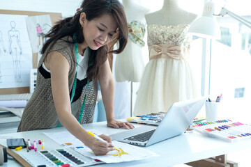 Happy Asian dressmaker or fashion designer uses laptop computer in her workshop, stylish woman in...