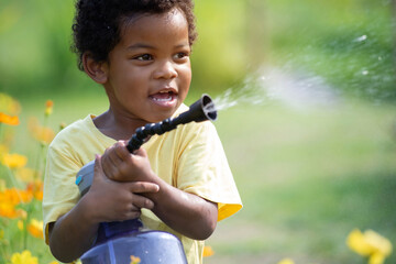 Happy African child boy is watering a flower bed with a large spray bottle, beautiful yellow flower...