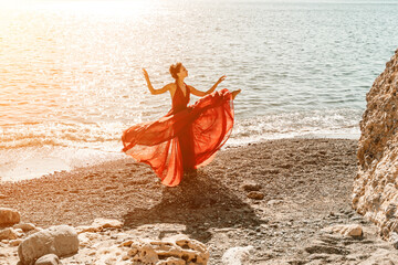 Woman red dress sea. Female dancer in a long red dress posing on a beach with rocks on sunny day. Girl on the nature on blue sky background. - obrazy, fototapety, plakaty