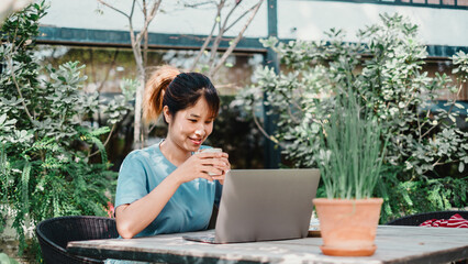 Woman in a blue top sipping coffee while using a laptop at a garden table surrounded by greenery.