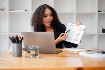 Smiling businesswoman points at a financial chart, engaging with content on her laptop at a wood-furnished office.