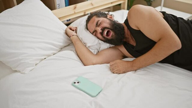 Hispanic man waking up, checking phone in a bright bedroom setting, portraying morning routine.