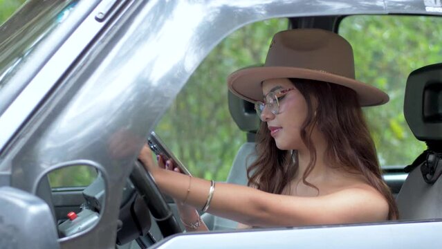 A woman in glasses and hat smiling while using her mobile phone in the driver's seat of a car.