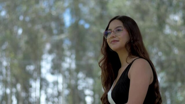 Elegant Hispanic woman with open hair and glasses enjoys a cheerful conversation in nature's embrace, surrounded by towering trees