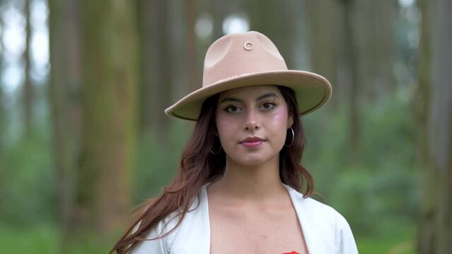 Beauty of an Ecuadorian woman in a hat and white top with open hair, as she finds tranquility in the forest, closing her eyes before slowly opening them with a radiant smile.