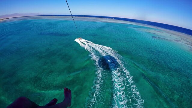 The downward view of a tourist parasailing behind a speedboat over an azure ocean.