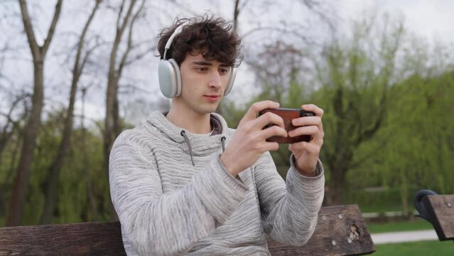 Male with curly hair and headphones use phone to play video game at outdoor park