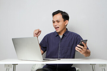 joyfull happy asian man holding mobile phone while sitting in front of laptop computer. on isolated background