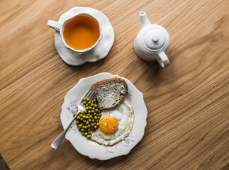 Delicious breakfast, brunch - fried egg, bread with butter, canned green peas and a cup of tea on a wooden background, top view - 786832901