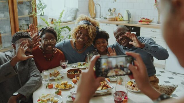 Happy African American family sitting together at dinner table, smiling and waving while posing for photo taken by girl with smartphone during home celebration