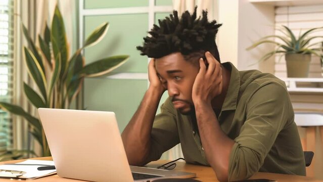 A man sitting at a laptop, head in hands, appears dejected and stressed, his expression gloomy. Houseplants and a window behind him suggest an indoor setting. 
