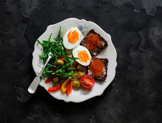 Delicious breakfast, brunch - fresh salad with arugula, olives, sweet pepper, boiled egg, red caviar sandwiches on a dark background, top view - 786834789