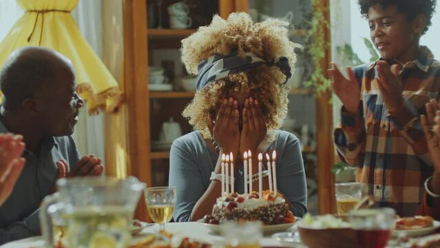 Little African American boy bringing birthday cake for mom covering her eyes with hands, she getting surprised, blowing candles and hugging kid as family clapping hands on home celebration