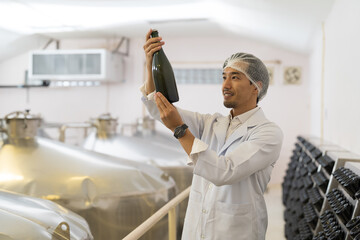 Male factory worker inspecting quality of wine bottles on shelf during manufacturing in process in...