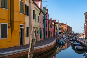 Blick auf die bunte Häuserfront in Burano (Italien), Boote schwimmen im Kanal, Häuser spiegeln sich im  Wasser - obrazy, fototapety, plakaty