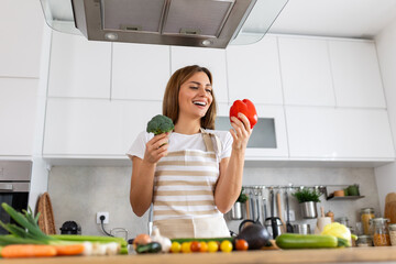 Photo of young woman smiling with red bell pepper while cooking and making salad with fresh vegetables in kitchen interior at home