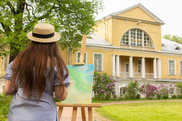 Female artist painting in spring lilac garden. Woman in hat with wooden easel and canvas standing...