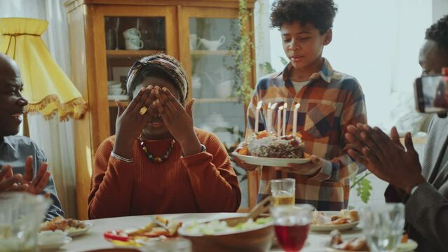 Young excited Black woman sitting with eyes closed, then blowing candles on cake and thanking family members during Birthday dinner party at home