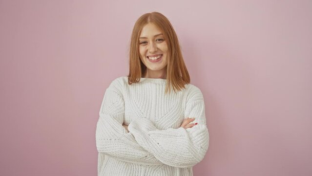 Confident redhead young woman in sweater, arms crossed, radiant smile on her face, standing against an isolated pink background, embodying positivity and success.