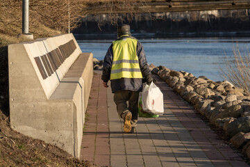 A man wearing a yellow vest walks down the promenade and checks the trash cans. homeless person. 