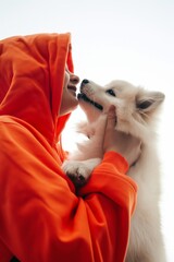 A person in an orange hoodie is affectionately touching noses with a white fluffy dog against a clear backdrop.