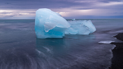 t&uuml;rkiser Eisbrocken (kleiner Eisberg) liegt auf schwarzem Lavastrand, Naturidyll Diamondbeach in Island, pastellfarbene Bew&ouml;lkung am fr&uuml;hen Morgen