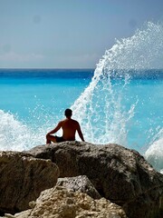 Man sitting on rocks and wave crashes in