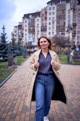 Fototapeta premium young adult woman in jeans and sand coat walks through the city on a spring day, modern architecture behind