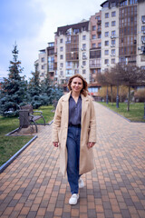 Fototapeta premium young adult woman in jeans and sand coat walks through the city on a spring day, modern architecture behind