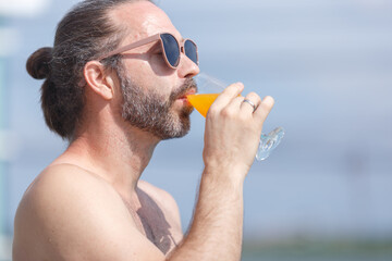 Very handsome man wear sunglass drinking orange juice walks along the poolside in summer.