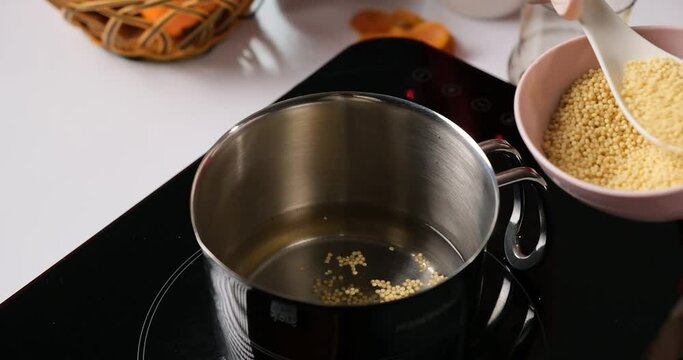 An image of a cooking process where a hand with a spoon pours small yellow millet pellets into a pot on an induction stove.