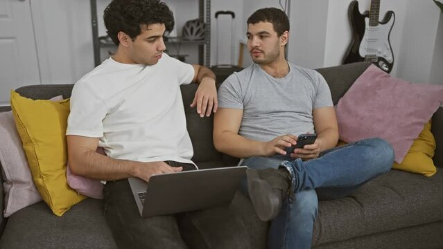 Two men casually using technology on a couch, one with a laptop, the other with a smartphone.