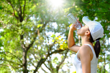 Female athlete lifts a bottle of drinking water in a clean bottle in hot weather with very sunny natural green background. Health and hydration concepts