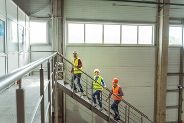 Factory workers, managers wearing hard hats, vests and work wear walking down stairs in warehouse