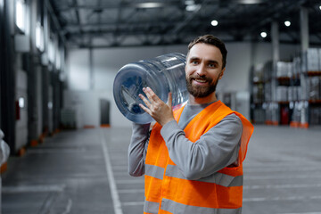 Attractive worker wearing vest and workwear carrying water bottle, looking at camera in warehouse