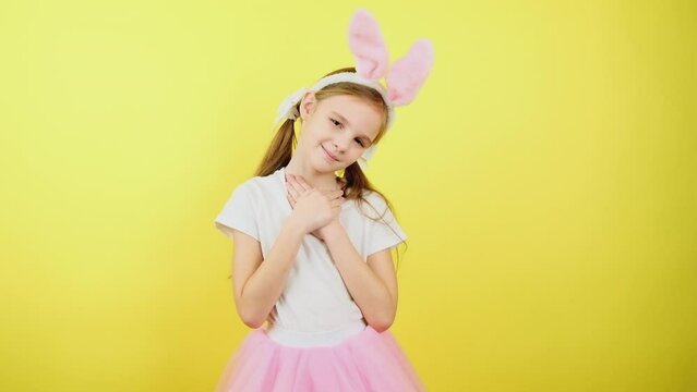 Happy easter! Portrait girl kid in pink bunny ears put folded palms on heart, isolated on yellow studio wall. Sign of sincere kind person, feeling thankful