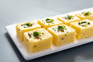 Yellow dhokla plated in a white plate on a grey background with negative space to write copy.