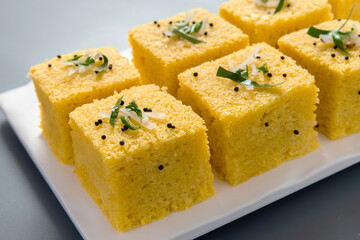 Yellow dhokla plated in a white plate on a grey background with negative space to write copy.