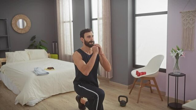 A bearded man in casual workout attire performs exercises in a modern bedroom, implying a home fitness routine.