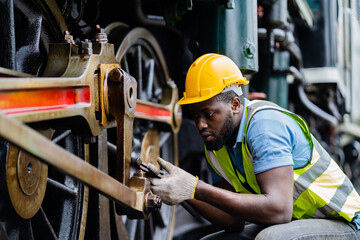 A man in a yellow helmet and safety vest is working on a train engine. He is wearing gloves and he...