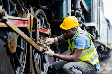 A man in a yellow helmet and safety vest is working on a train engine. He is wearing gloves and he...