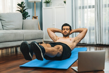 Athletic and sporty man doing crunch on fitness mat while follow online home workout exercise...