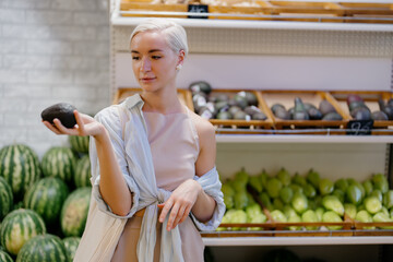 A young female shopper examines an avocado with a thoughtful expression in the produce section of a...