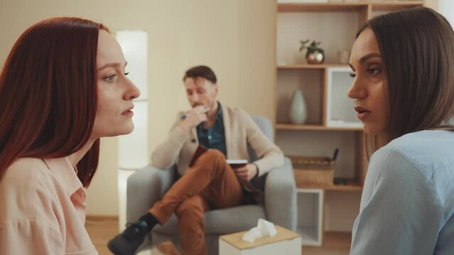 Lesbian couple deeply engaged in a therapy session, articulating their points while a therapist observes, facilitating their path to understanding