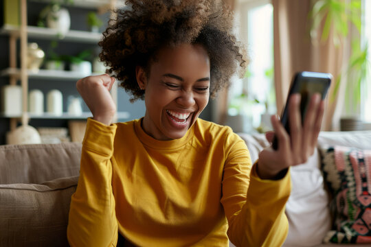 An excited young Afro-American woman holds a smartphone while sitting on a sofa at home. Her joyful expression reflects happiness and satisfaction. She looks at the mobile smartphone screen