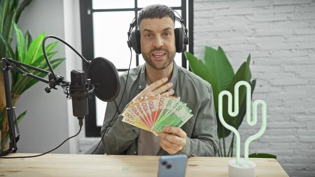 Bearded man in a radio studio examining norwegian kroner bills with headphones and microphone in front of him.