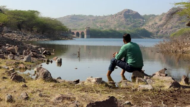 isolated young man sitting at stone at pristine lake shore at morning