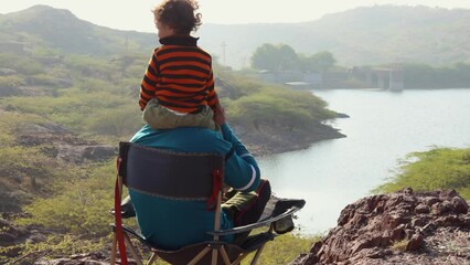 son and father sitting at camping chair at mountain top with lake view at morning