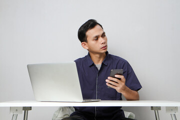 pensive confused asian man holding mobile phone whiile sitting in front of laptop computer. isolated background